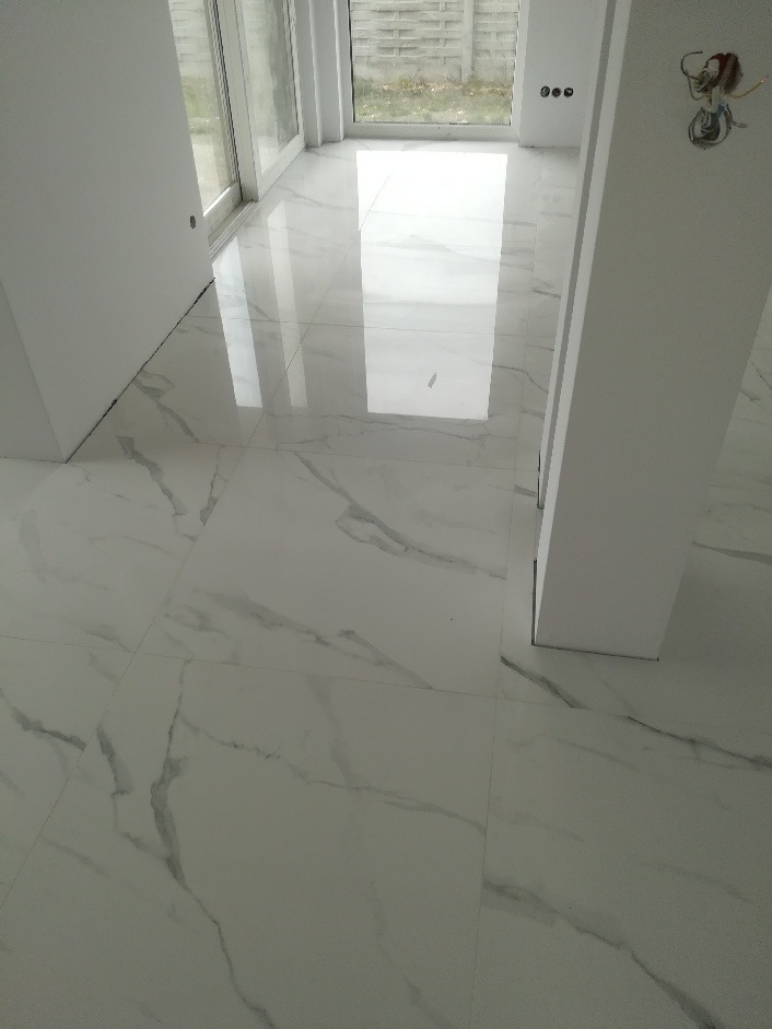 Białe, polerowane płytki podłogowe z szarym marmurkowym wzorem, ułożone w jasnym pomieszczeniu z dużymi oknami i widoczną instalacją elektryczną na ścianie.