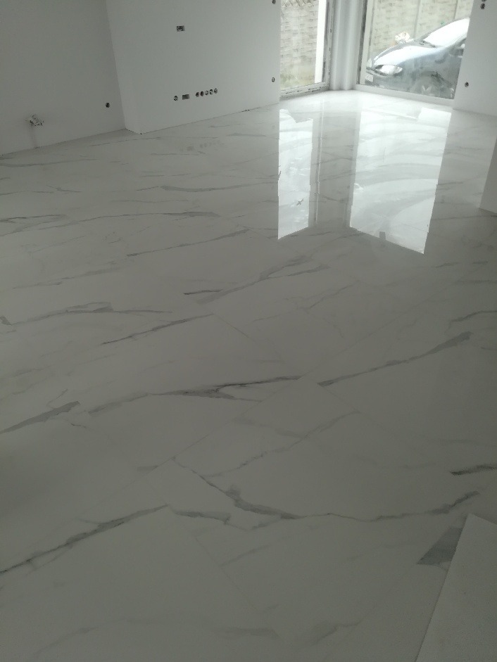 Błyszcząca, biała podłoga z marmurowym wzorem w pustym, jasnym pomieszczeniu z widocznym oknem i odbijającym się w podłodze światłem.