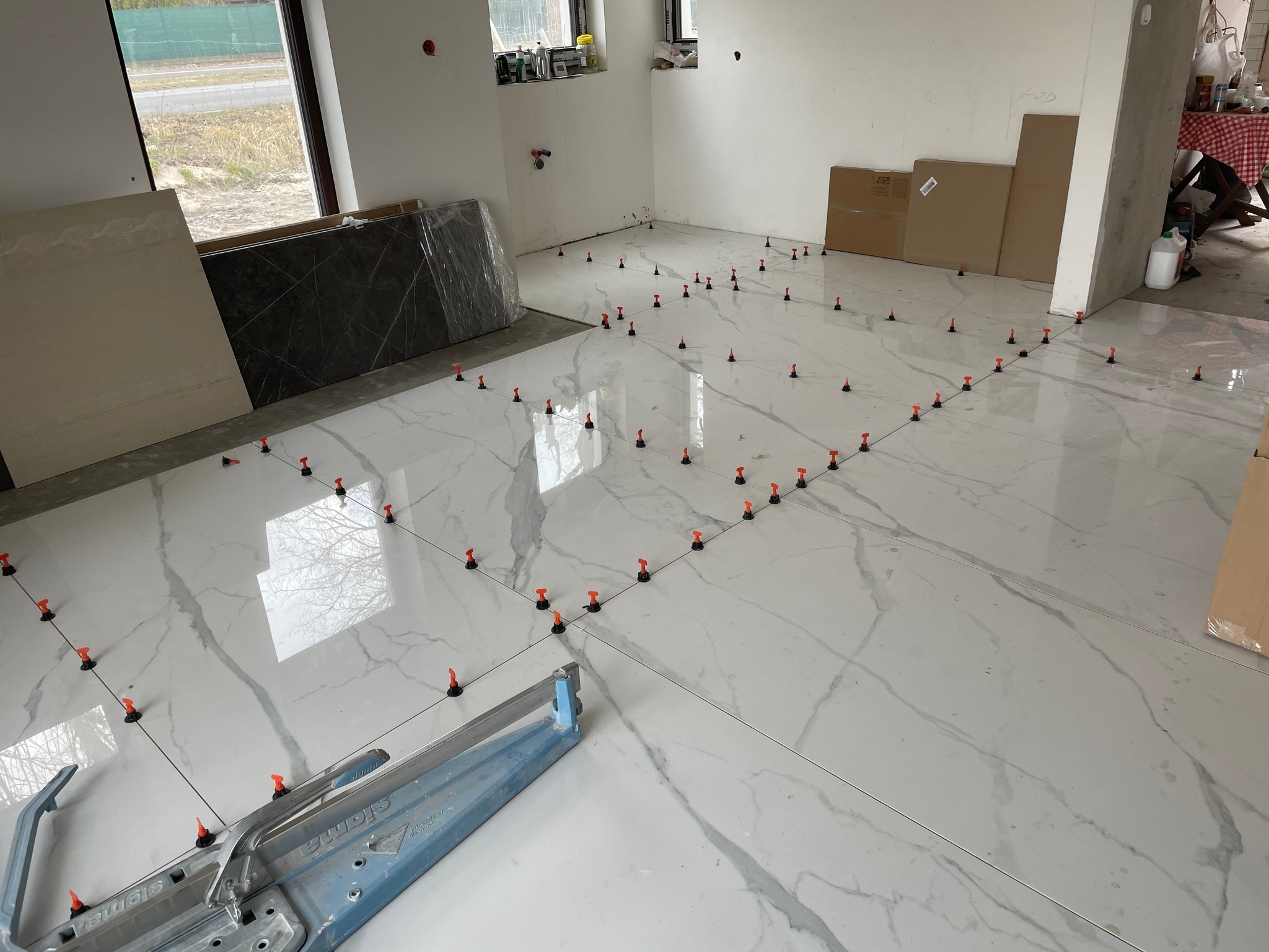 Układanie białych płytek podłogowych z marmurkowym wzorem, z użyciem poziomujących klipsów i maszynki do cięcia płytek Sigma, w trakcie remontu wnętrza.