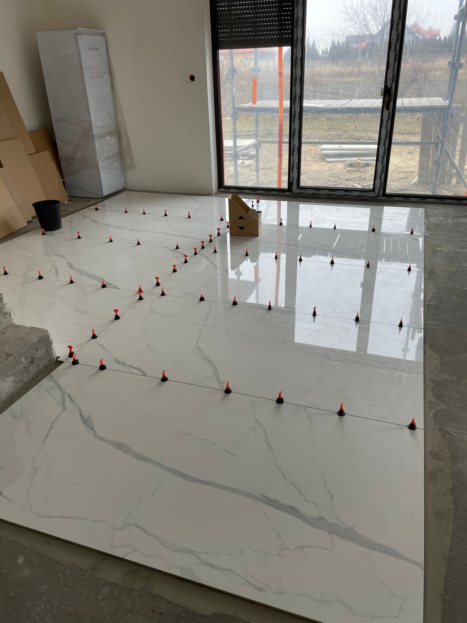 Układanie płytek podłogowych z wzorem marmuru, z użyciem systemu poziomowania z czerwonymi klipsami, widok na okno z rusztowaniami na zewnątrz.
