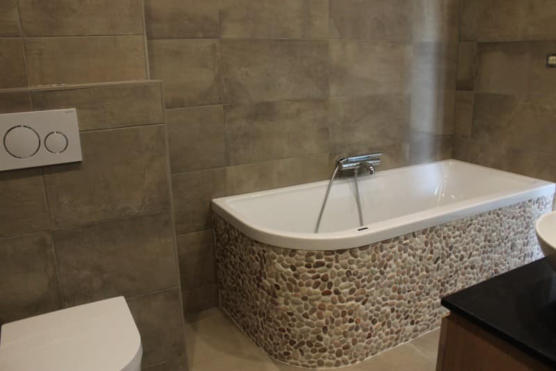 Nowoczesna łazienka z wanną obłożoną kamieniami rzecznymi, szare płytki na ścianach i podwieszana toaleta z białym przyciskiem spłukującym.