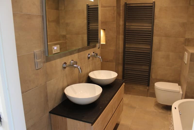 Nowoczesna łazienka z dwoma umywalkami nablatowymi, bateria ścienna chromowana, lustro z podświetleniem, grzejnik drabinkowy w kolorze grafitowym i beżowe płytki imitujące kamień.