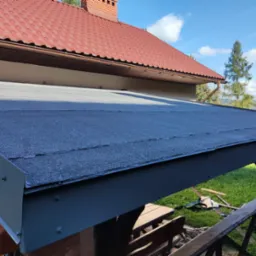 Widok na dach altany pokryty papą bitumiczną, z widocznym fragmentem dachu domu pokrytego czerwoną dachówką ceramiczną w tle, oraz narzędzia dekarza leżące na trawie.