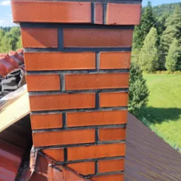 Ceglany komin z czarną fugą, wystający ponad czerwoną dachówkę, na tle zieleni drzew i łąk.