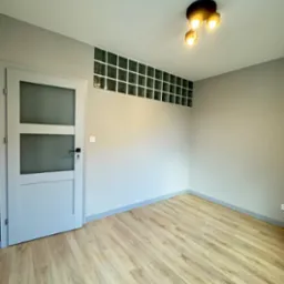 Pusty, świeżo wykończony pokój z panelami podłogowymi, szarą ścianą, białymi drzwiami z szybą i dekoracyjnym pasem z luksferów pod sufitem, oświetlony trzypunktową lampą sufitową.