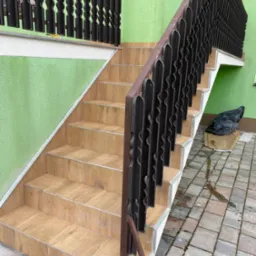 Wyłożone płytkami schody zewnętrzne z brązową balustradą o dekoracyjnym wzorze, prowadzące do wejścia z zieloną ścianą. Na podeście widoczny czarny worek na śmieci.