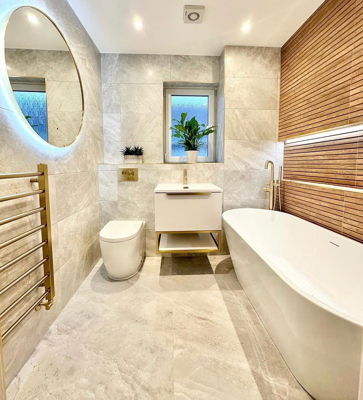 Elegancka łazienka z marmurowymi płytkami, owalnym lustrem z podświetleniem LED, białym wolnostojącym zlewem z złotymi akcentami, wiszącą toaletą i drewnianą ścianą z oświetleniem punktowym.