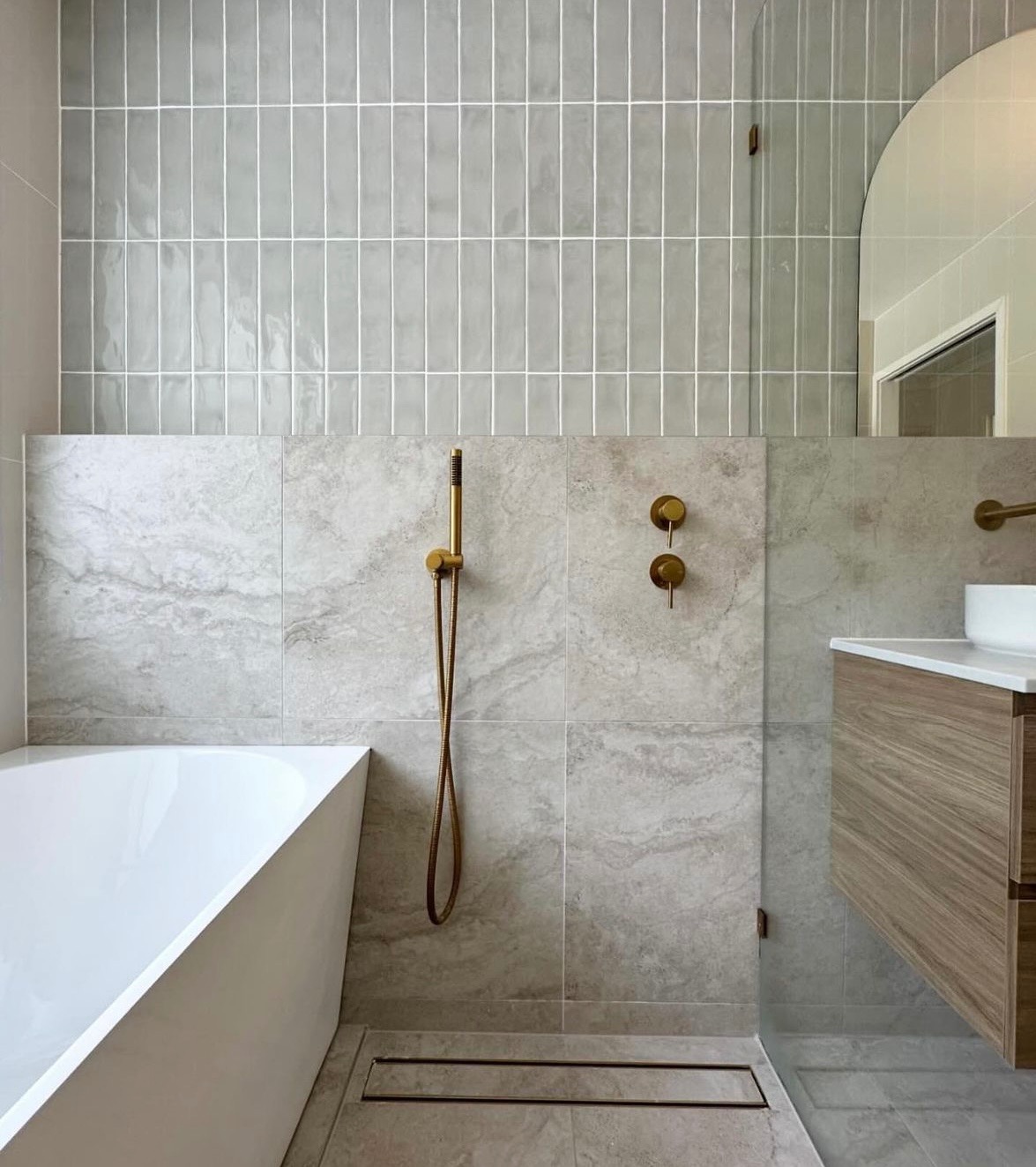 Nowoczesna łazienka z szarymi płytkami imitującymi kamień, złotą armaturą, minimalistyczną kabiną prysznicową typu walk-in i drewnianą szafką pod umywalką.