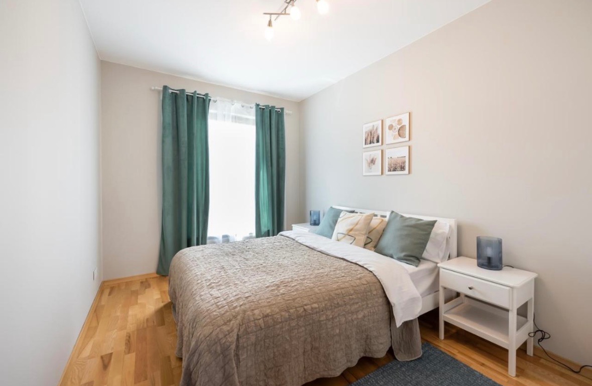 Jasna sypialnia z drewnianą podłogą, białym łóżkiem i szafkami nocnymi, zielonymi zasłonami i dekoracjami ściennymi z motywami roślinnymi.