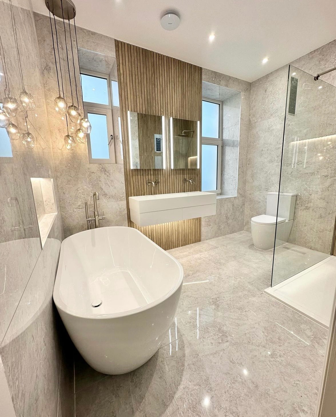 Nowoczesna łazienka z wolnostojącą wanną, podwójną umywalką z podświetleniem LED, drewnianą ścianką dekoracyjną i kabiną prysznicową ze szklaną ścianą. Zwisające lampy kuliste dodają elegancji.