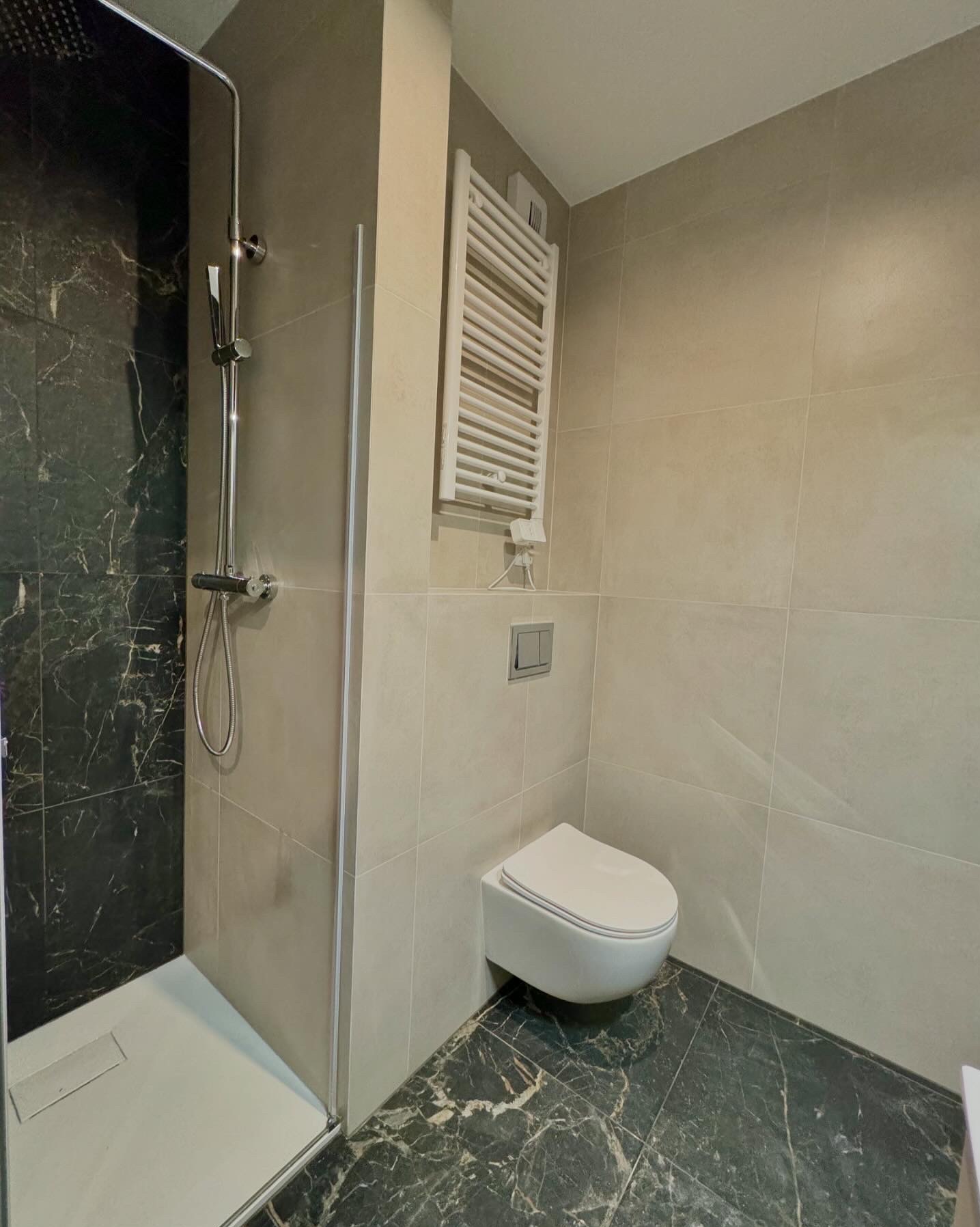 Nowoczesna łazienka z kabiną prysznicową typu walk-in, marmurowymi płytkami na podłodze i ścianie prysznica, oraz podwieszaną toaletą i grzejnikiem drabinkowym.