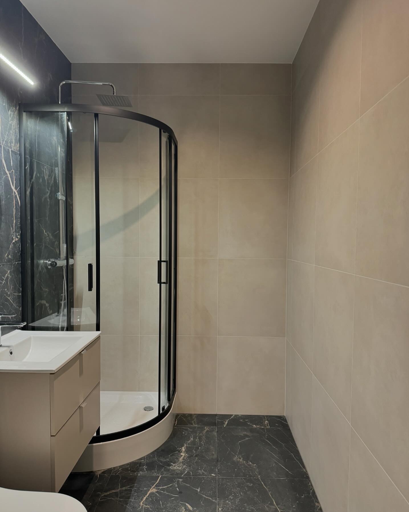 Nowoczesna łazienka z czarną kabiną prysznicową typu walk-in, umywalką w beżowej szafce i marmurowymi płytkami na podłodze i jednej ścianie.