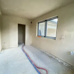 Wnętrze pomieszczenia po nałożeniu tynków maszynowych, widoczne instalacje grzewcze podłogowe w kolorze czerwonym i niebieskim oraz okno z widokiem na budynek w trakcie budowy.