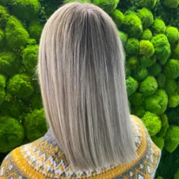 Widok z tyłu na świeżo ostrzyżone i pofarbowane włosy w odcieniach blondu, z delikatnym ombre, na tle ściany z mchu i wzorzystego swetra.