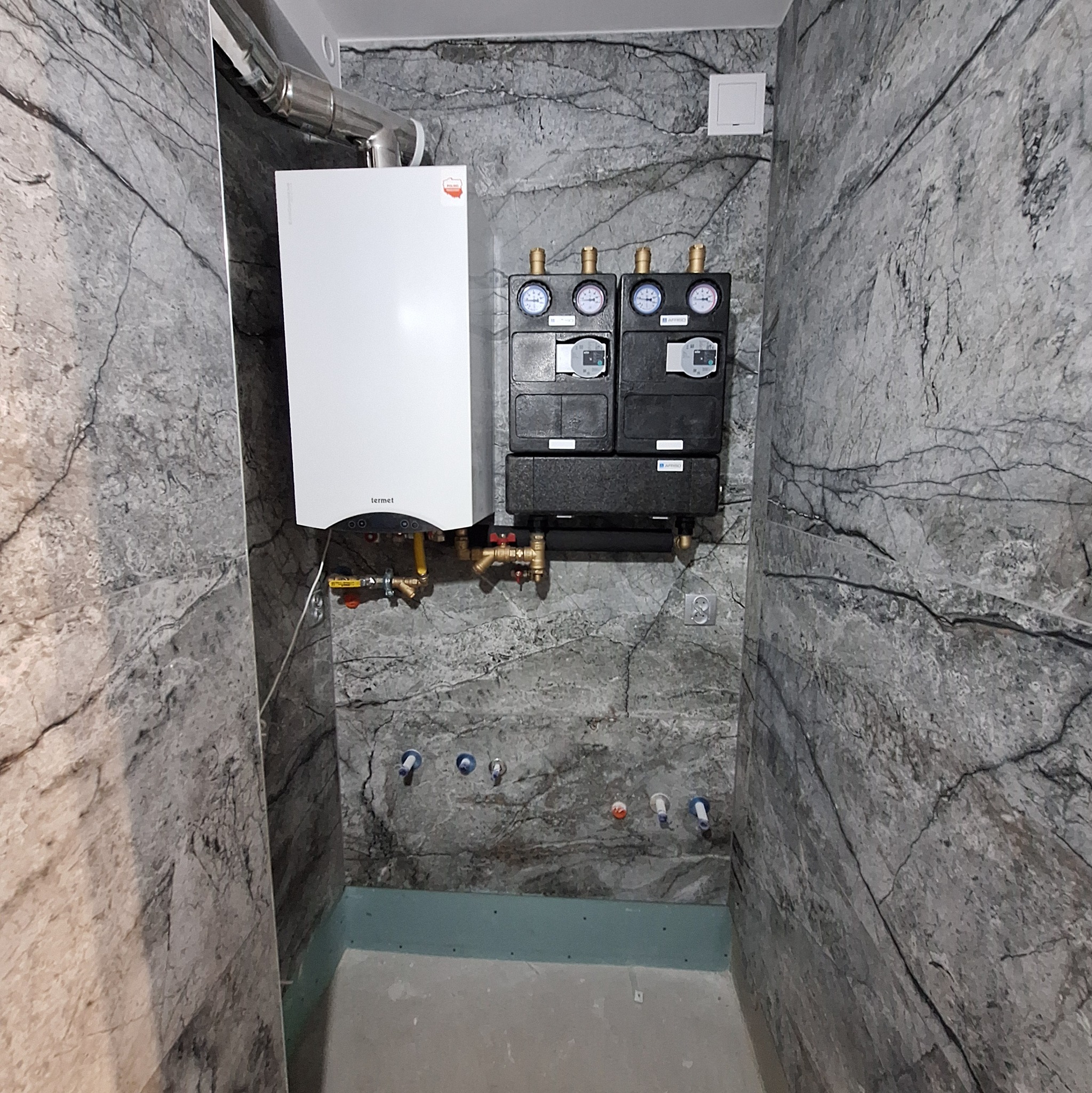 Biały piec gazowy marki Termet zamontowany na ścianie wyłożonej płytkami imitującymi marmur, z widocznymi czarnymi rozdzielaczami i rurami instalacji centralnego ogrzewania.