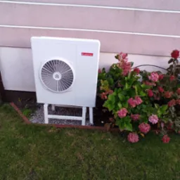 Biała jednostka zewnętrzna z logo Ariston, zamontowana na metalowym stelażu na żwirowym podłożu, obok rabaty z kwitnącymi hortensjami i trawnikiem.