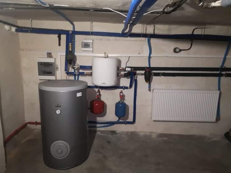 Instalacja grzewcza w piwnicy w Nakle nad Notecią, widoczny duży szary zbiornik, mały biały zbiornik, grzejnik panelowy i niebieskie rury.