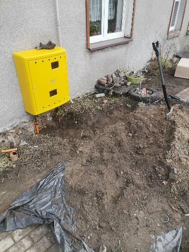 Żółta skrzynka na ścianie budynku w Nakle nad Notecią, obok wykop pod instalację z widoczną łopatą i folią zabezpieczającą.