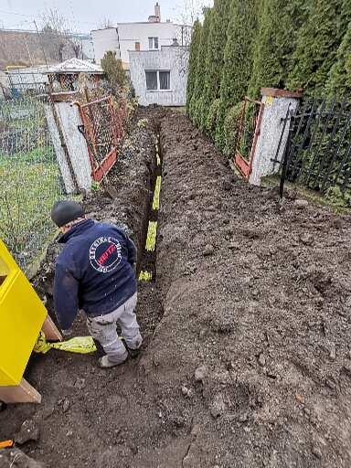Wykop w ziemi z widocznymi żółtymi rurami, instalator w roboczym ubraniu pochylony nad wykopem, w tle budynek mieszkalny i ogrodzenie.