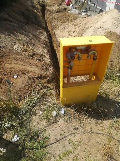 Żółta skrzynka z zaworami i rurami na zewnątrz, obok wykopu w ziemi, prawdopodobnie w trakcie instalacji.