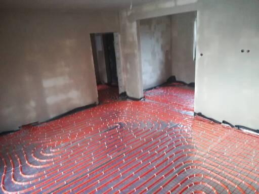 Czerwone rurki układane spiralnie na podłodze z siatki, przygotowane do zalania wylewką w surowym wnętrzu z widocznymi otworami drzwiowymi.