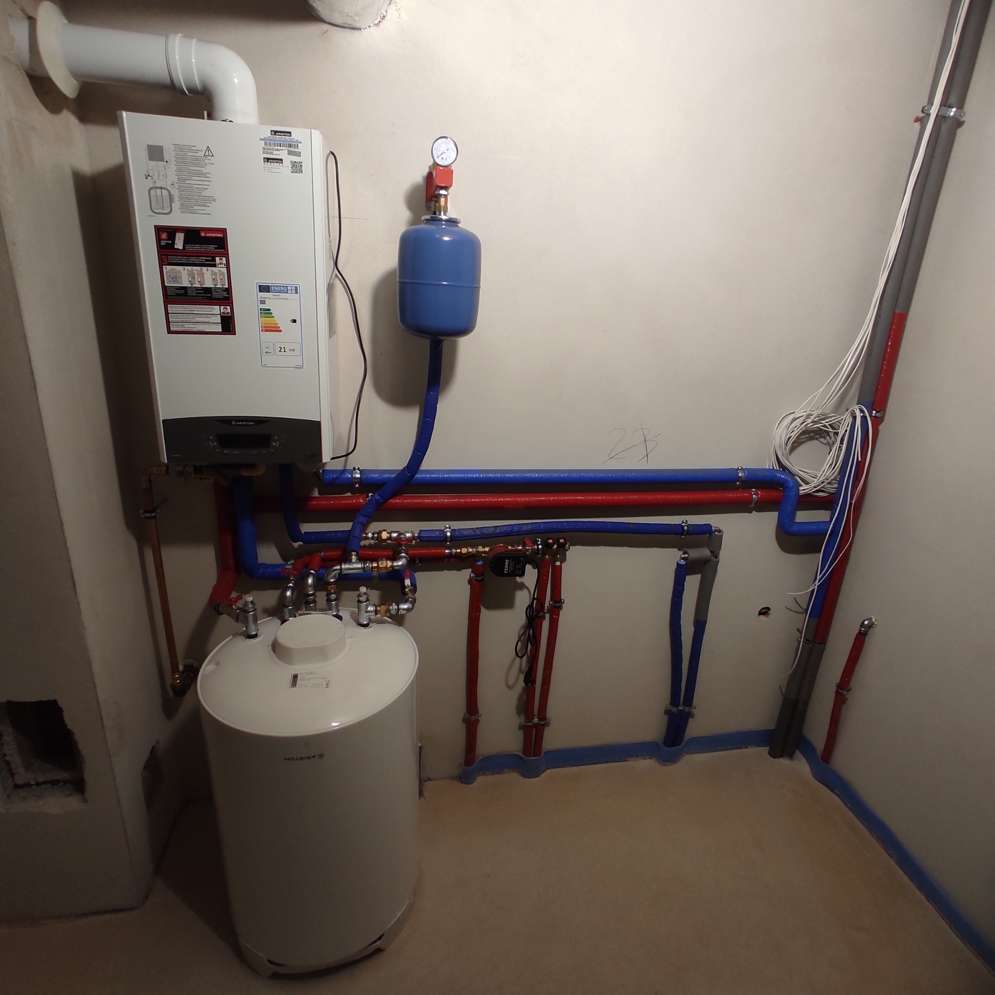 Biały piec gazowy Ariston z podłączonymi rurami w kolorze czerwonym i niebieskim, zbiornik wyrównawczy, instalacja w surowym stanie.