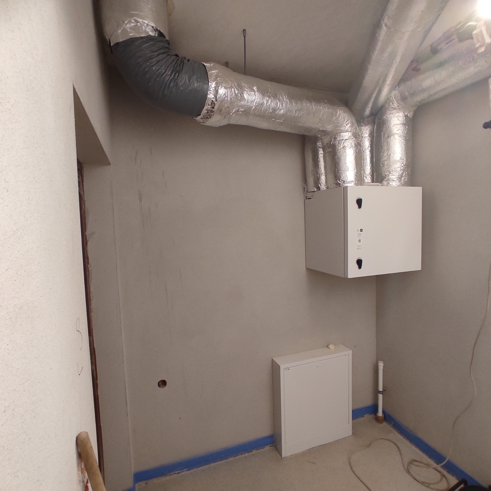 Biała centrala wentylacyjna z podłączonymi, zaizolowanymi przewodami wentylacyjnymi w surowym wnętrzu, widoczna również biała skrzynka na ścianie i zabezpieczenia podłogi.