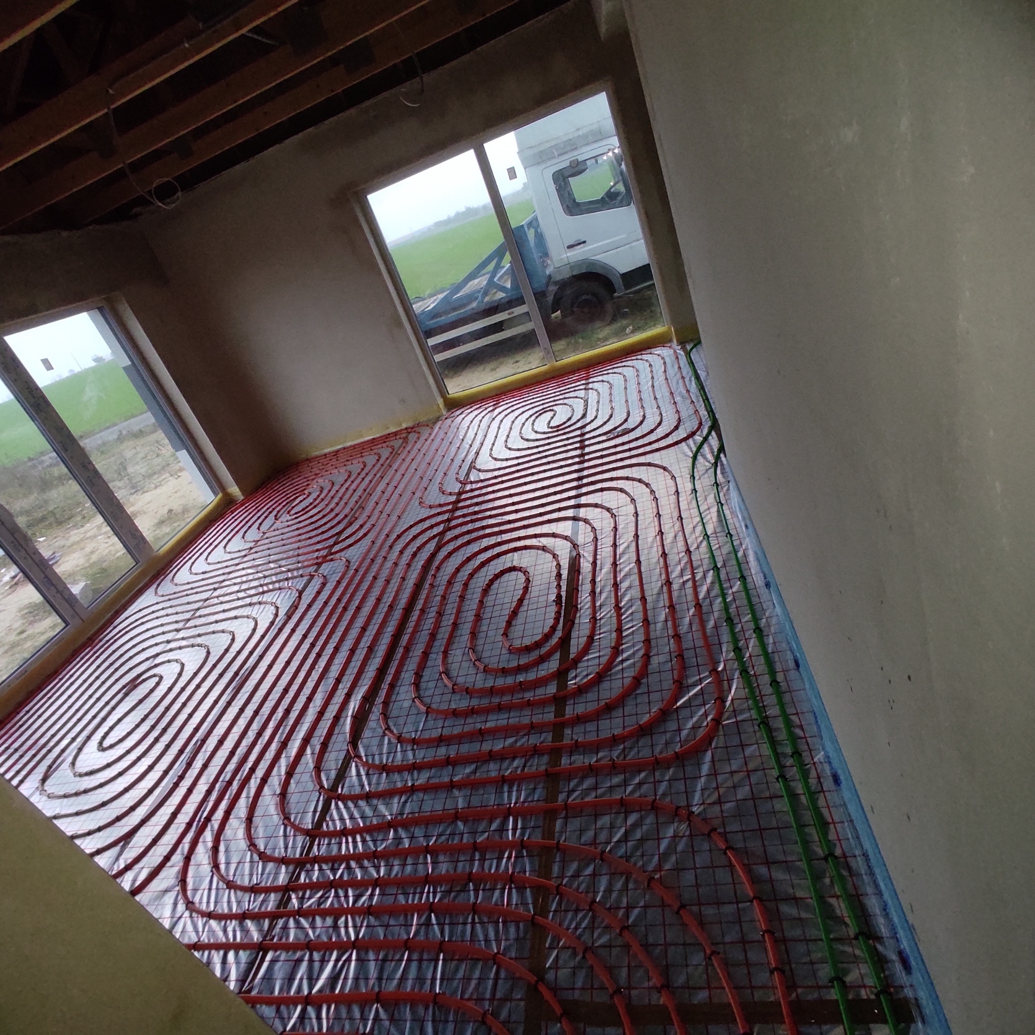 Wnętrze w surowym stanie z widocznym systemem rur ogrzewania podłogowego ułożonym w spiralne wzory, na tle okna z widokiem na ciężarówkę na zewnątrz.
