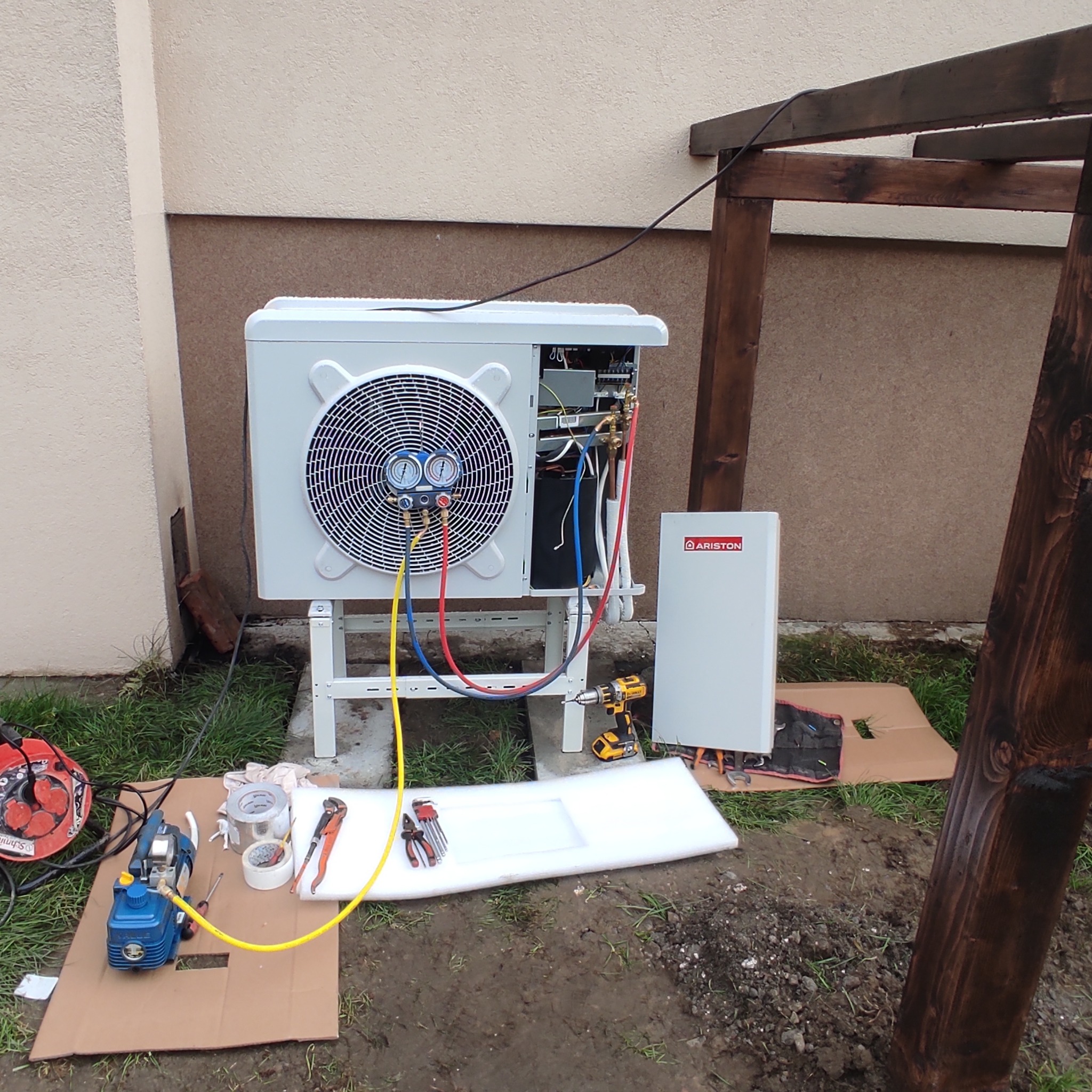 Biała jednostka zewnętrzna z podłączonymi manometrami, obok biała jednostka wewnętrzna marki Ariston, narzędzia i pompa próżniowa na kartonie, tło stanowi ściana budynku i drewniana konstrukcja.