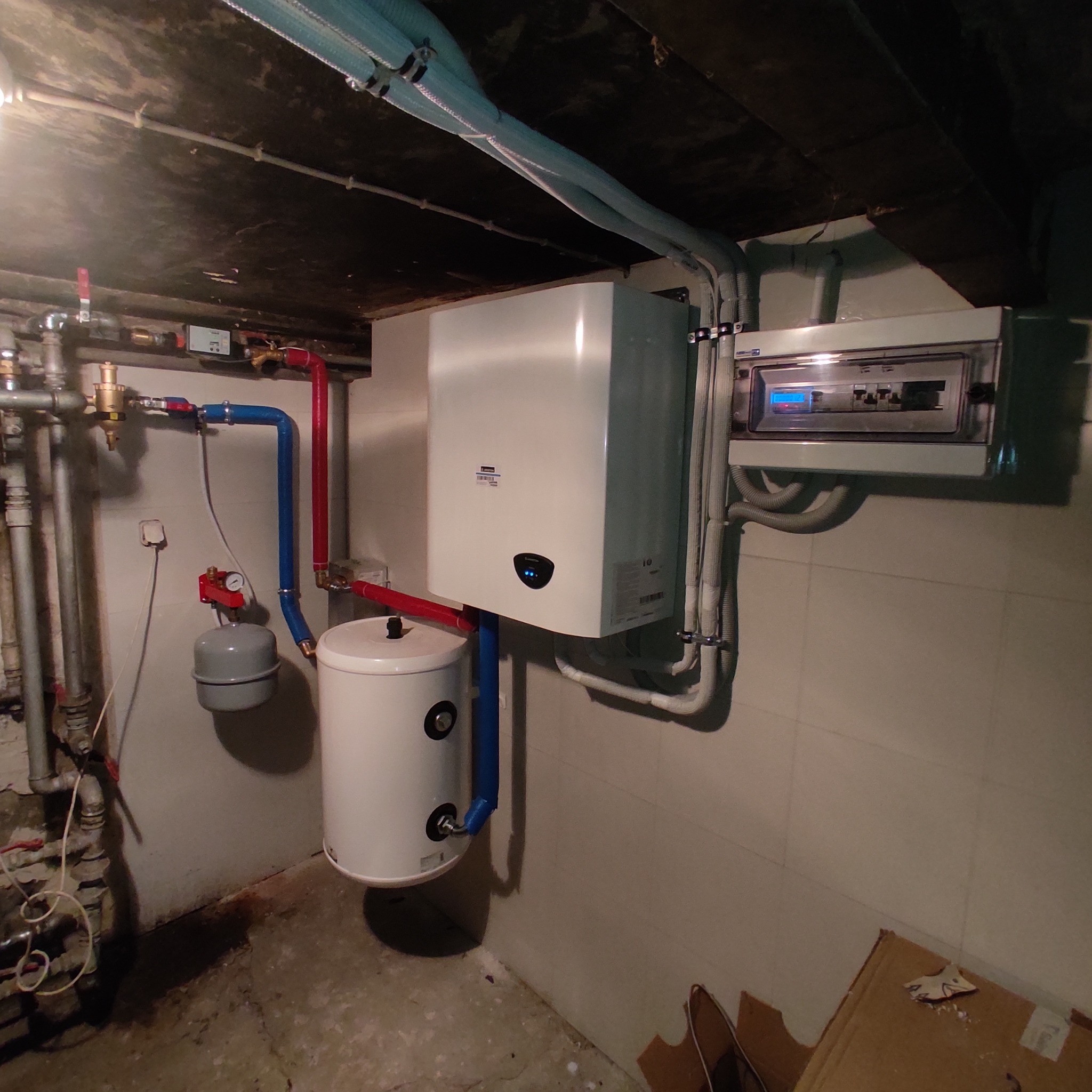 Zestaw urządzeń grzewczych w piwnicy: wiszący kocioł kondensacyjny z podłączonym zasobnikiem ciepłej wody, zabezpieczenia elektryczne i instalacja hydrauliczna z rurami w izolacji, widoczne złącza...