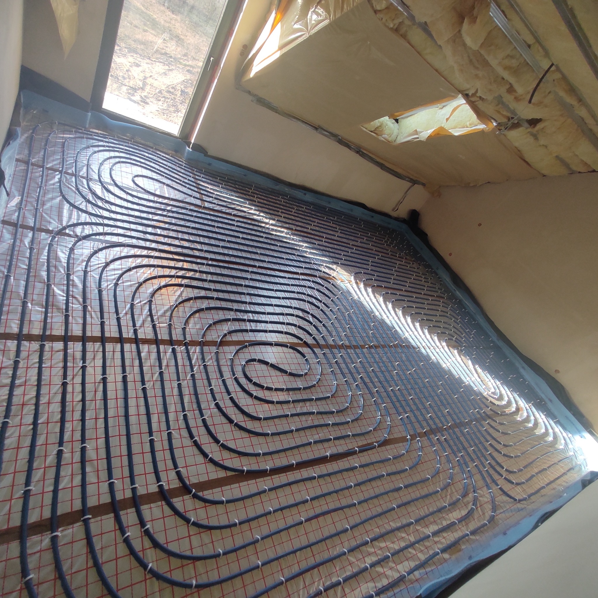 Montaż rur ogrzewania podłogowego na folii z siatką w nowym pomieszczeniu z oknem dachowym i izolacją ścian.