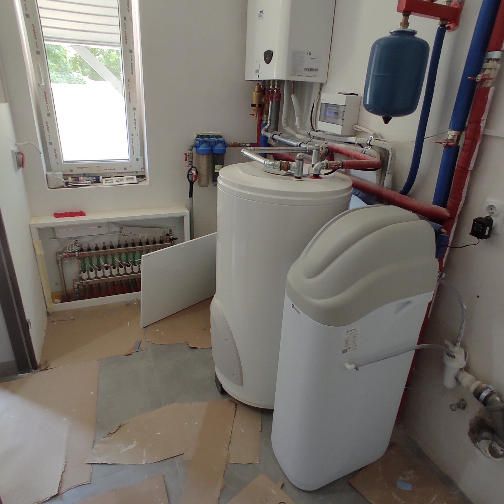 Wnętrze kotłowni z zamontowanym zasobnikiem ciepłej wody użytkowej, zmiękczaczem wody i rozdzielaczem ogrzewania podłogowego w szafce pod oknem.