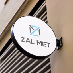 Okrągły szyld firmy ŻAL-MET z niebiesko-szarym logo przymocowany do beżowej ściany budynku z widoczną kratką wentylacyjną.