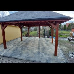 Mykola Bilous - Zadaszona wiata z drewnianymi słupami i szarym dachem, wyłożona szarą kostką brukową, widok z zewnątrz na tle ogrodu z wózkiem budowlanym