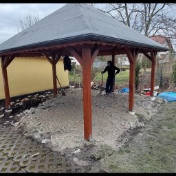 Mykola Bilous - Drewniana altana z ciemnym dachem w trakcie budowy, widoczny mężczyzna z łopatą przygotowujący podłoże pod kostkę, częściowo ułożona kostka brukowa na obrzeżach, rozkopana ziemia wewnątrz konstrukcji.