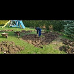 Mykola Bilous - Pracownik w ciemnej bluzie i niebieskich spodniach kopie ziemię łopatą na trawniku, wyznaczony kwadrat z wbitymi palikami, obok taczki i narzędzia, w tle zielony żywopłot i niebieski plac zabaw.