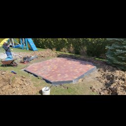 Mykola Bilous - Ośmiokątna powierzchnia wyłożona czerwoną kostką brukową z czarnym obrzeżem, widoczna na trawniku w ogrodzie, obok leży zagęszczarka, taczka i wiadro, w tle plac zabaw dla dzieci.