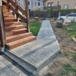 Ułożona ścieżka z szarej kostki brukowej prowadząca od schodów wejściowych do domu, z zamontowanym odpływem liniowym, widok na posesję z trawnikiem i częściowo ułożoną kostką.