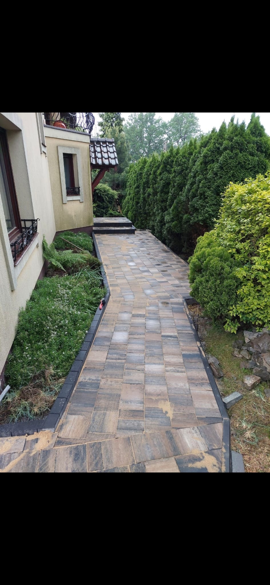 Ścieżka z kostki brukowej prowadząca wzdłuż domu, z widocznymi obrzeżami i roślinnością, częściowo zasypana piaskiem.