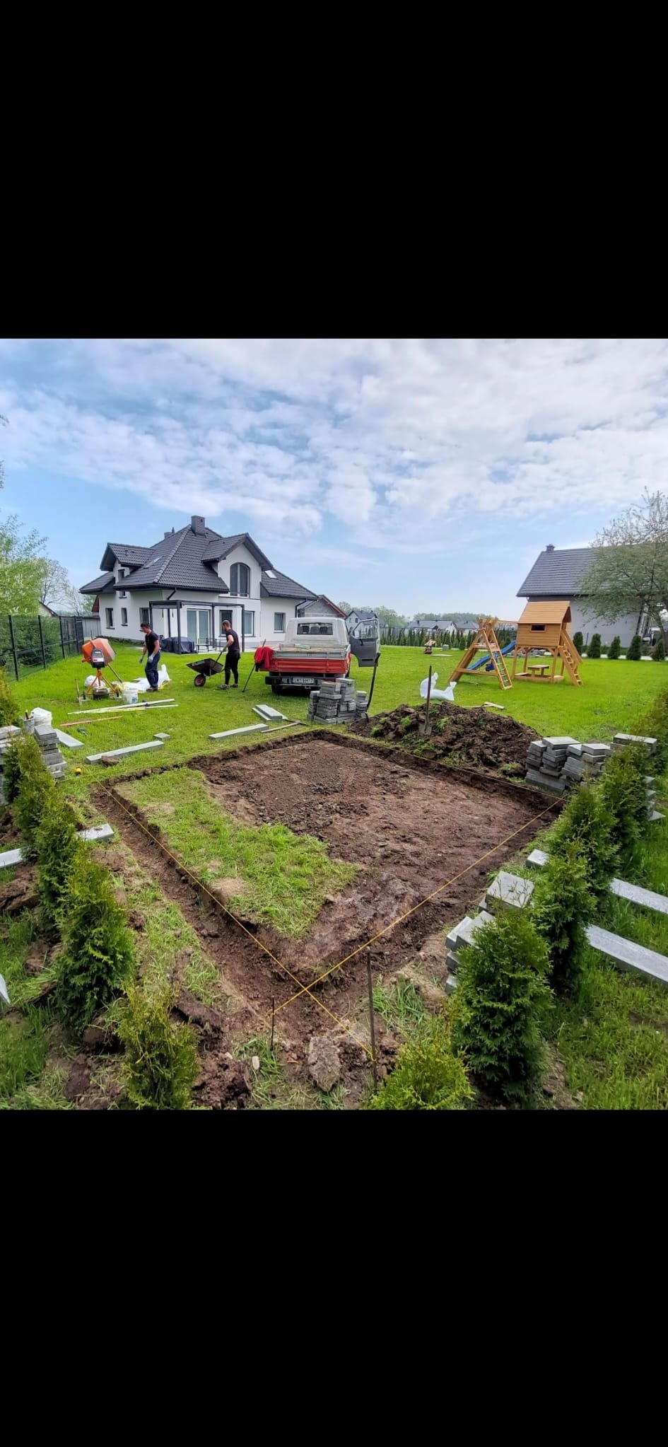 Przygotowanie podłoża pod kostkę brukową: wytyczony obszar z rozciągniętymi sznurkami, obok ułożone kostki, w tle ekipa z taczką i betoniarką oraz samochód dostawczy na tle domu jednorodzinnego...