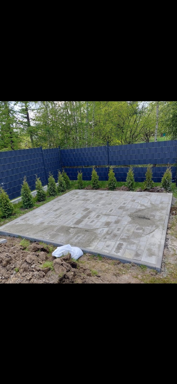 Przygotowany plac pod taras z szarych kostek, obrzeża wykończone, częściowo zasypany piaskiem, w tle rząd małych tuj i ciemny płot panelowy.