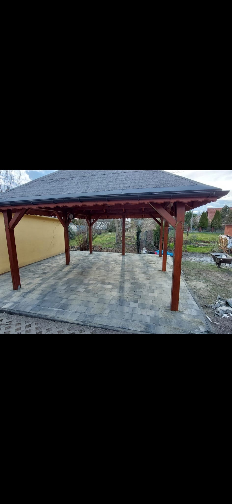 Zadaszona wiata z drewnianymi słupami i szarym dachem, wyłożona szarą kostką brukową, widok z zewnątrz na tle ogrodu z wózkiem budowlanym