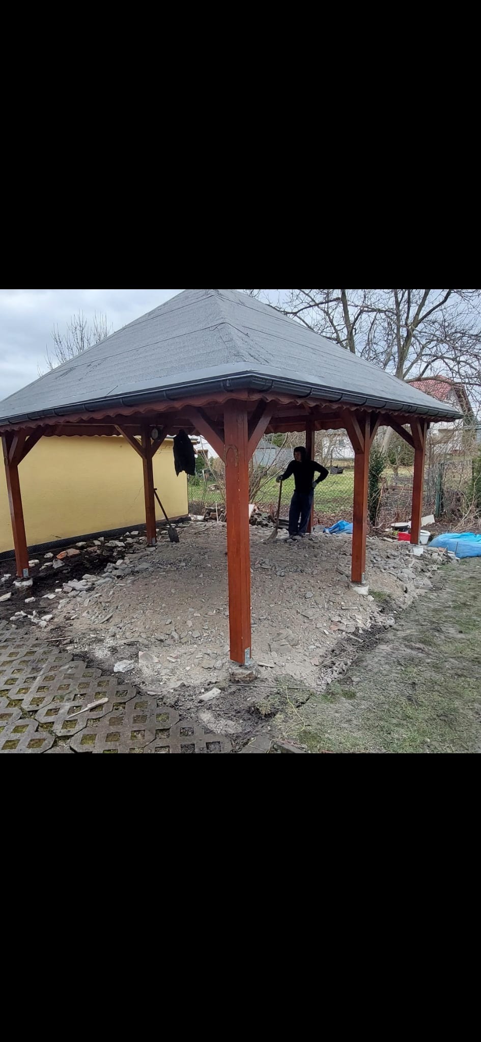 Drewniana altana z ciemnym dachem w trakcie budowy, widoczny mężczyzna z łopatą przygotowujący podłoże pod kostkę, częściowo ułożona kostka brukowa na obrzeżach, rozkopana ziemia wewnątrz konstrukcji.