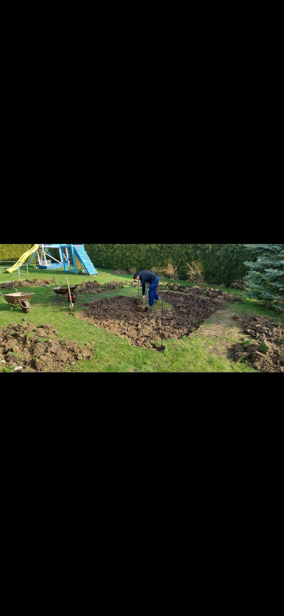 Pracownik w ciemnej bluzie i niebieskich spodniach kopie ziemię łopatą na trawniku, wyznaczony kwadrat z wbitymi palikami, obok taczki i narzędzia, w tle zielony żywopłot i niebieski plac zabaw.