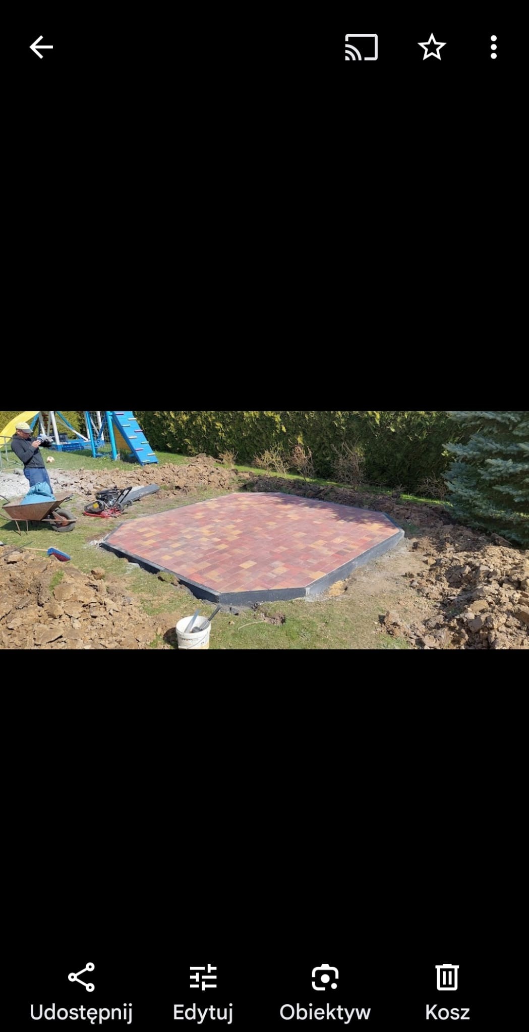 Ośmiokątna powierzchnia wyłożona czerwoną kostką brukową z czarnym obrzeżem, widoczna na trawniku w ogrodzie, obok leży zagęszczarka, taczka i wiadro, w tle plac zabaw dla dzieci.