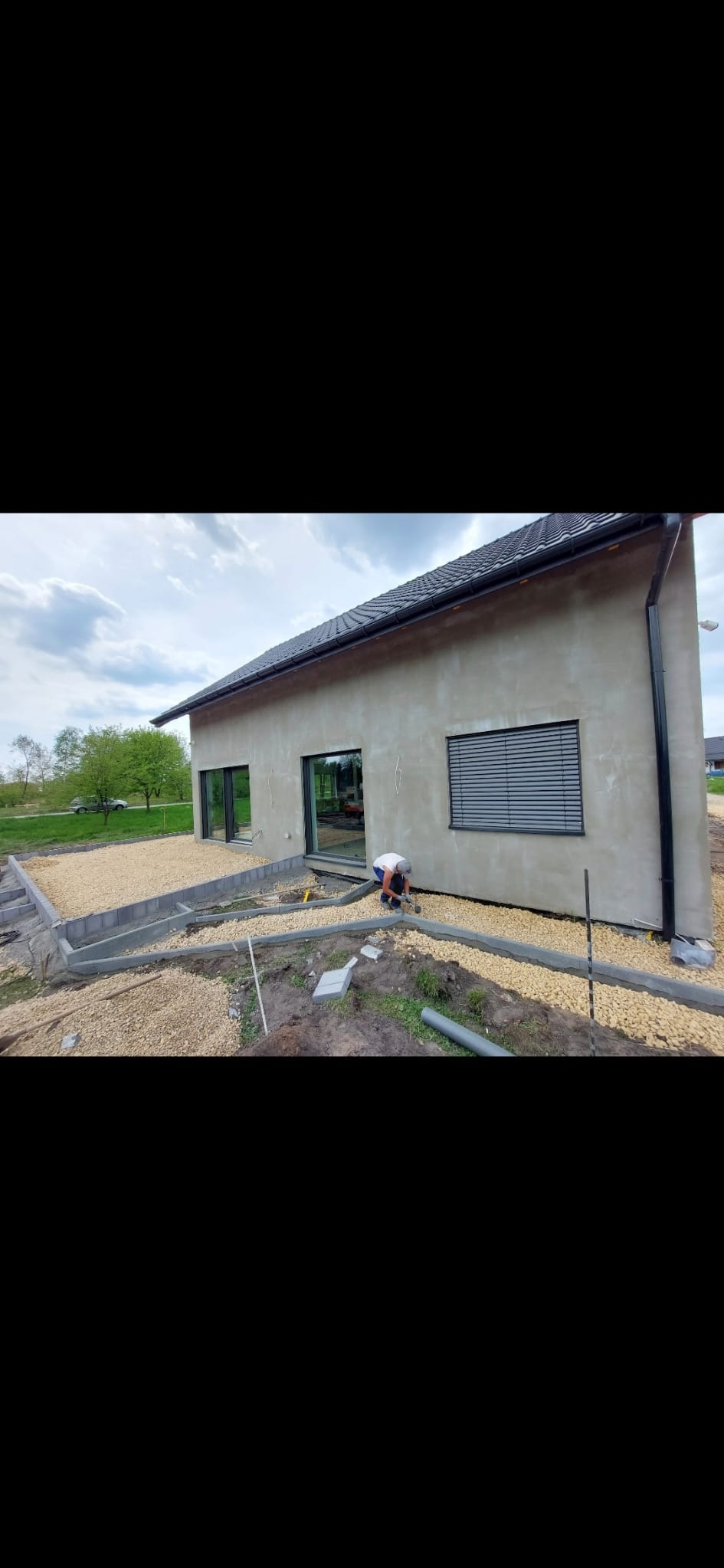 Pracownik w trakcie układania kamieni ozdobnych wokół nowo wybudowanego domu z szarą elewacją, widoczne obrzeża betonowe i narzędzia pracy.