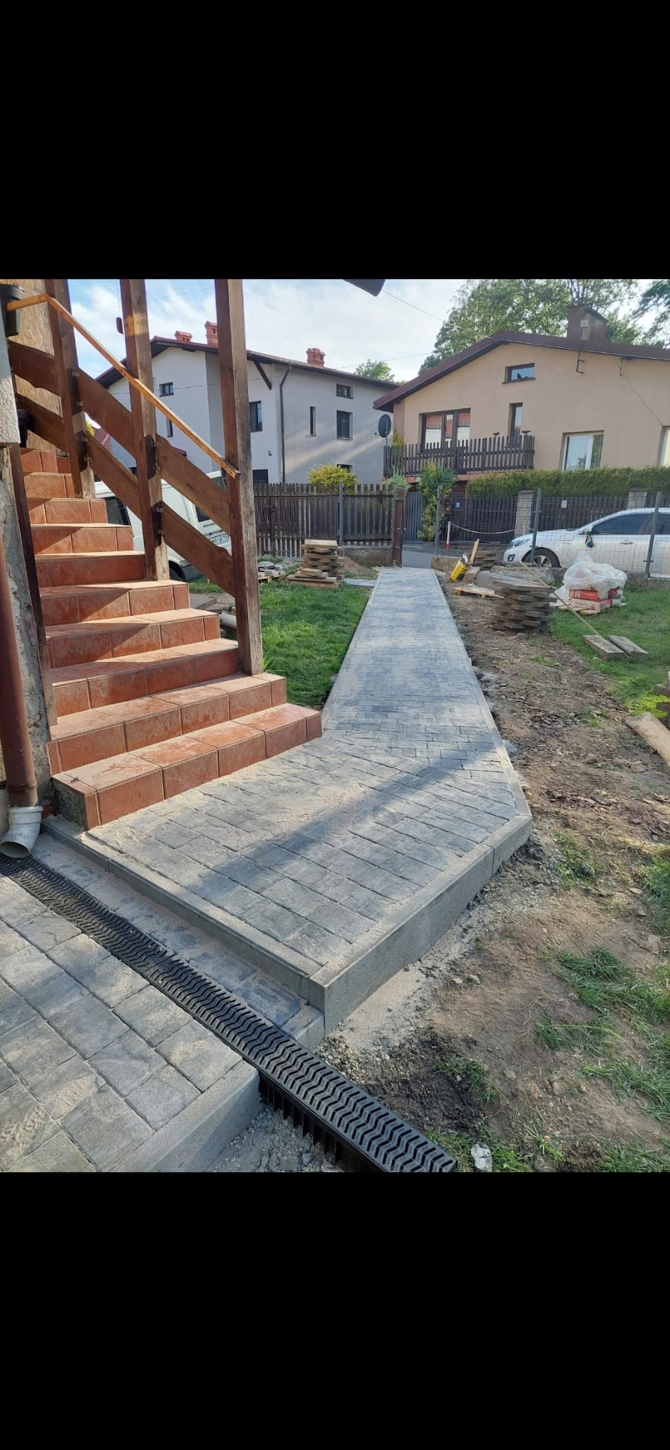 Ułożona ścieżka z szarej kostki brukowej prowadząca od schodów wejściowych do domu, z zamontowanym odpływem liniowym, widok na posesję z trawnikiem i częściowo ułożoną kostką.