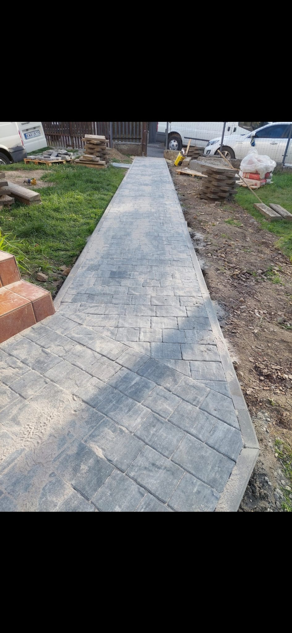 Świeżo ułożony chodnik z szarej kostki brukowej o nieregularnym kształcie, z widocznymi fugami i pozostałościami piasku, prowadzący wzdłuż trawnika i ogrodzenia, z zaparkowanymi samochodami w tle.