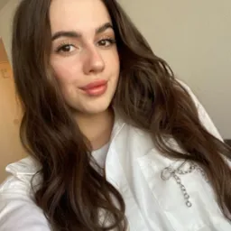 Portret kobiety z długimi brązowymi włosami, ubrana w białą koszulę z ozdobną srebrną broszką na kieszeni.