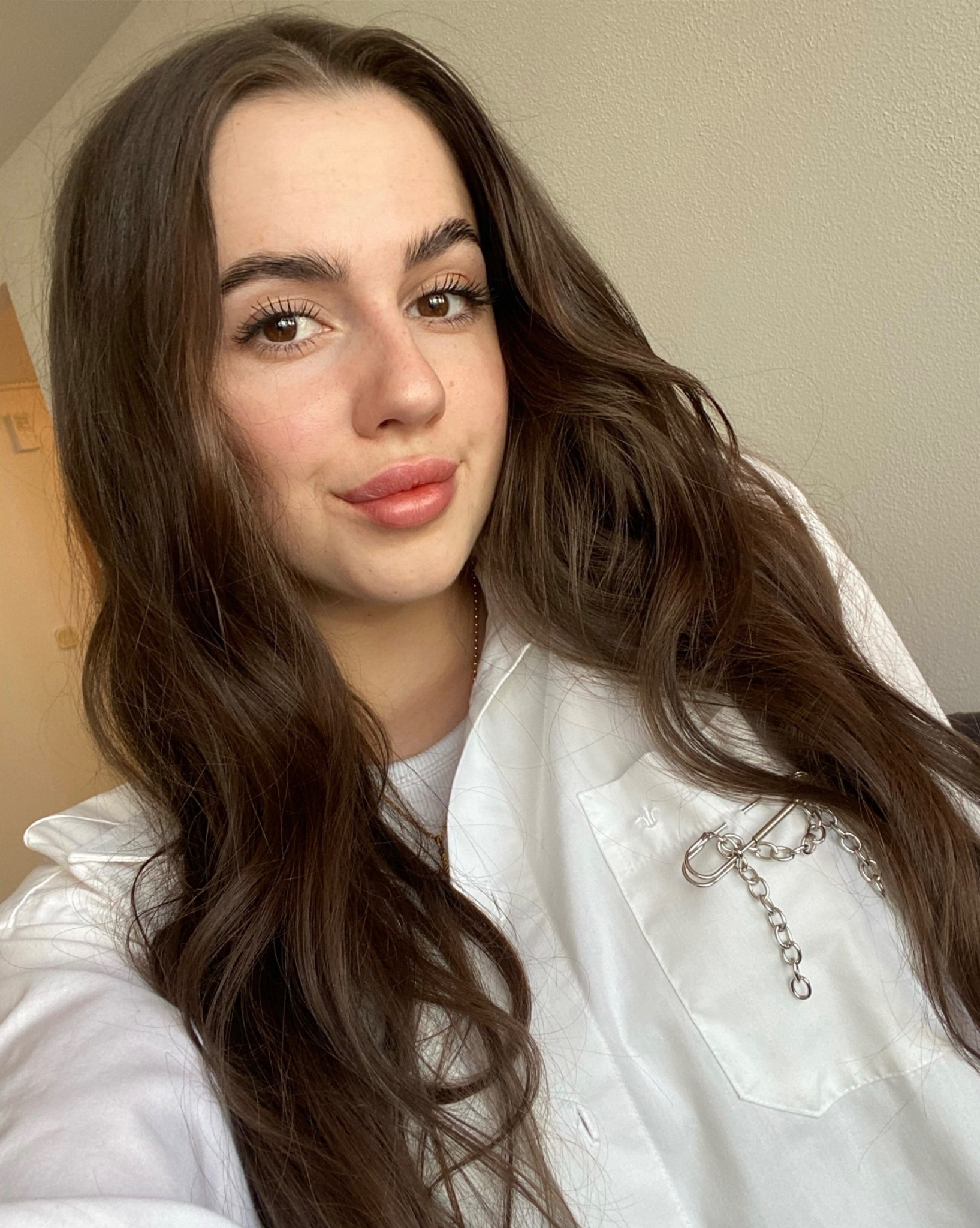 Portret kobiety z długimi brązowymi włosami, ubrana w białą koszulę z ozdobną srebrną broszką na kieszeni.